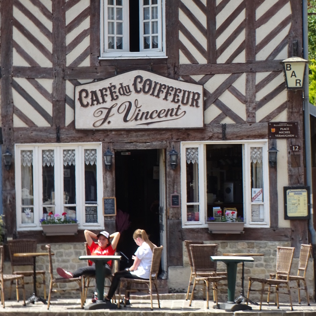 Deux jeunes se reposent sur une terrasse devant le 'Café du Coiffeur', un batiment à colombage à Beuvron-en-Auge