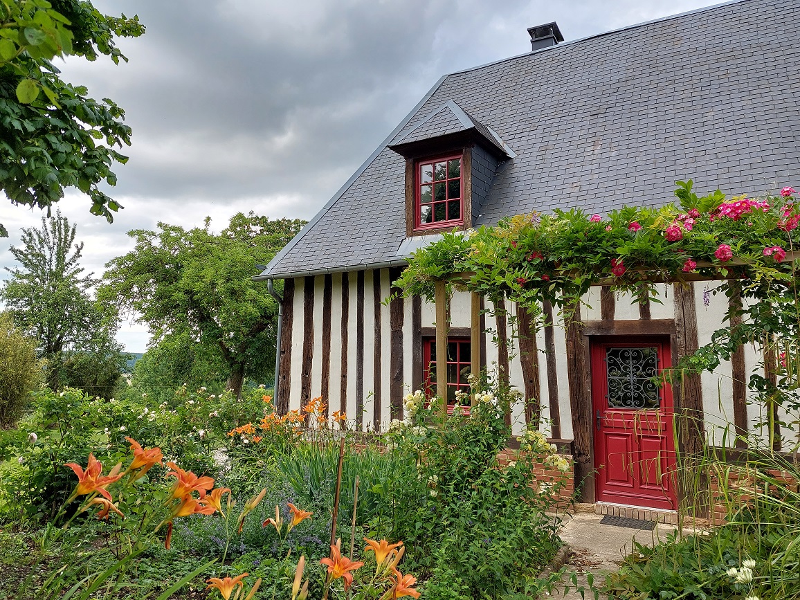 Image de l'exterieur d'une maison à colombages, avec une porte et des fenetres peintes en rouge, avec un jardin fleuri en premier plan