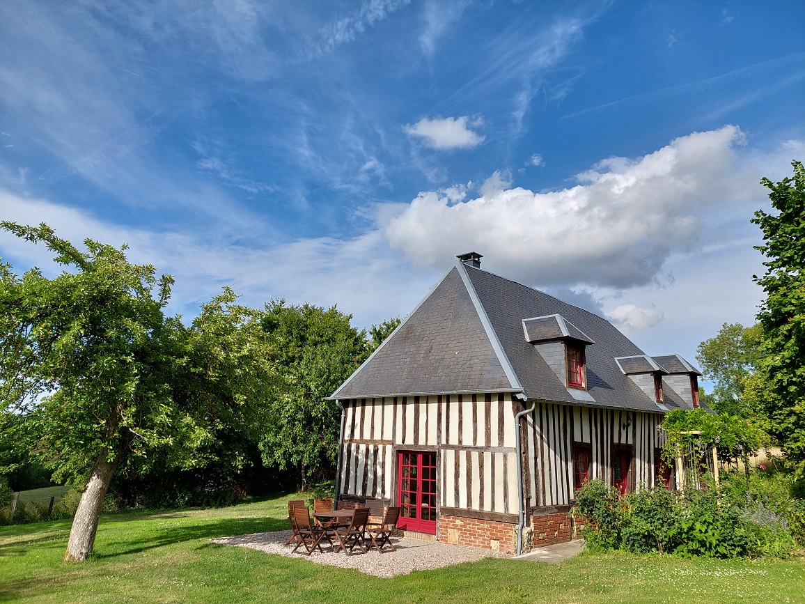 Image d'une maison à colombages entourée d'herbe et des arbres verts, le tout sous un ciel bleu avec quelques nuages blanches